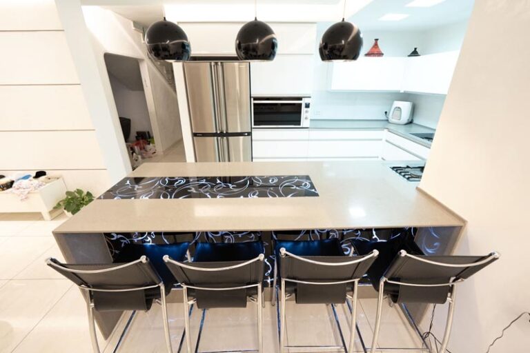 Front view of kitchen island bar with four black stools, decorative panels, dome pendants, and stainless refrigerator behind - modern kitchen