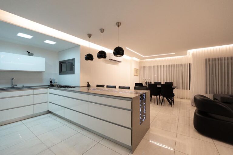 Handleless white kitchen island under black pendant lights with LED cove ceiling and dining area beyond - modern kitchen remodel in Houston, TX