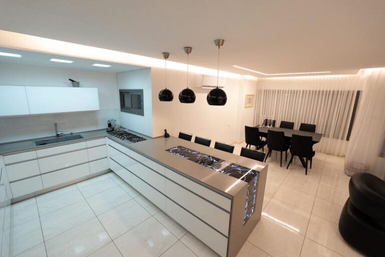 Wide view of white modern kitchen with island cooktop, black dome pendant lights, and dining table in the open-plan layout - modern kitchen