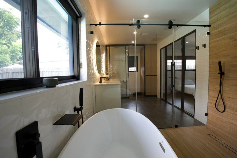 Freestanding tub beside window with black barn-door glass shower enclosure and glass partition view into bedroom - condo bathroom remodeling in