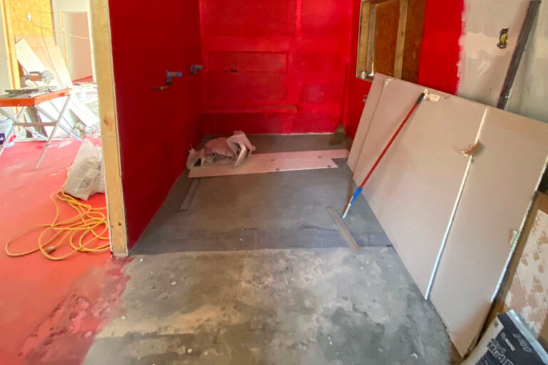 Shower area mid-remodel with red liquid waterproofing membrane applied to walls over bare concrete subfloor - condo bathroom remodeling in
