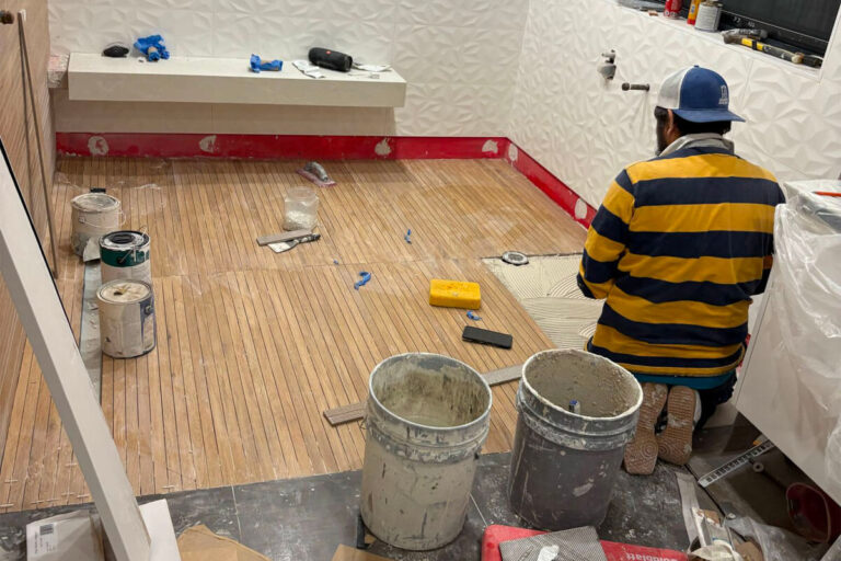 Tile setter laying wood-look porcelain floor planks with thinset buckets, red waterproofing visible at wall base - condo bathroom remodeling in